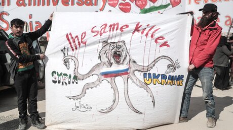 Protestteilnehmer in Idlib, Syrien, während einer Demonstration gegen das syrische Regime am 15.03.22, dem 11. Jahrestag des Beginns des syrischen Bürgerkriegs. Auf dem Plakat ist ein Bär mit der Russlandflagge zu sehen sowie die Schriftzüge: Syrien, Ukraine und Der selbe Mörder. / © Anas Alkharboutli (dpa)