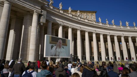 Menschen beobachten Papst Franziskus auf einem riesigen Monitor, der auf dem Petersplatz aufgestellt ist, wie er die Gläubigen segnet. / © Andrew Medichini (dpa)