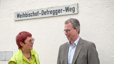Historikerin Marita Krauss (l.) und Rainer Schnitzler, Bürgermeister von Pöcking, stehen unter dem Straßenschild des Weihbischof-Defregger-Wegs. / © Christoph Renzikowski (KNA)