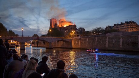 Menschen stehen am Ufer der Seine und blicken auf den Brand der Kathedrale Notre-Dame in Paris am 15. April 2019. / © Corinne Simon (KNA)