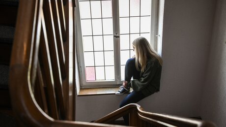 Eine junge Frau sitzt einsam am Fenster  / © Julia Steinbrecht (KNA)