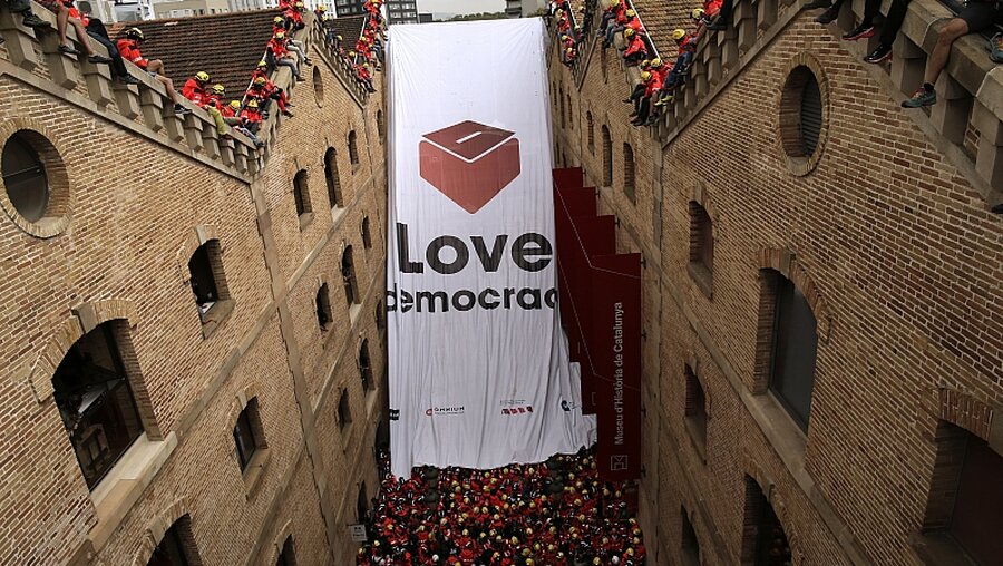 Katalanische Feuerwehrleute entfalten ein riesiges Banner mit einer Wahlurne / © Manu Fernandez (dpa)