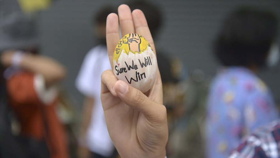Ein Demonstrant zeigt den Drei-Finger-Gruß als Zeichen des Widerstands und hält dabei ein Osterei mit der Aufschrift "Sure we will win" (Wir werden sicher gewinnen) / © Uncredited/AP/ (dpa)