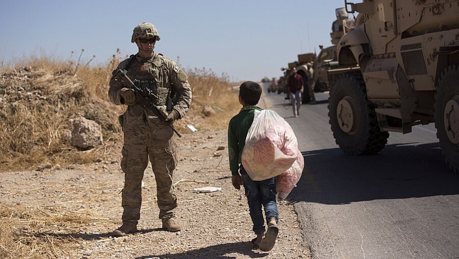 Ein syrischer Junge, der Süßigkeiten verkauft, geht an einem US-Soldaten vorbei, während der ersten gemeinsamen Bodenpatrouille in der sogenannten Sicherheitszone auf der syrischen Grenze zur Türkei / © Maya Alleruzzo (dpa)