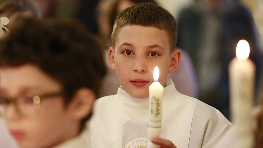 Ein Junge trägt bei seiner Kommunion eine weiße Kommunionalbe und hält eine brennende Kommunionkerze in der Hand.  / © Natalia Gileva (KNA)
