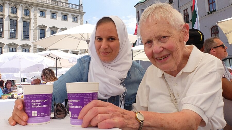 Fatima Zohra Medjadi (l.) und Marianne Rogall sitzen an der Friedestafel während des Hohen Friedensfestes vor dem Augsburger Rathaus.  / © Christopher Beschnitt (KNA)