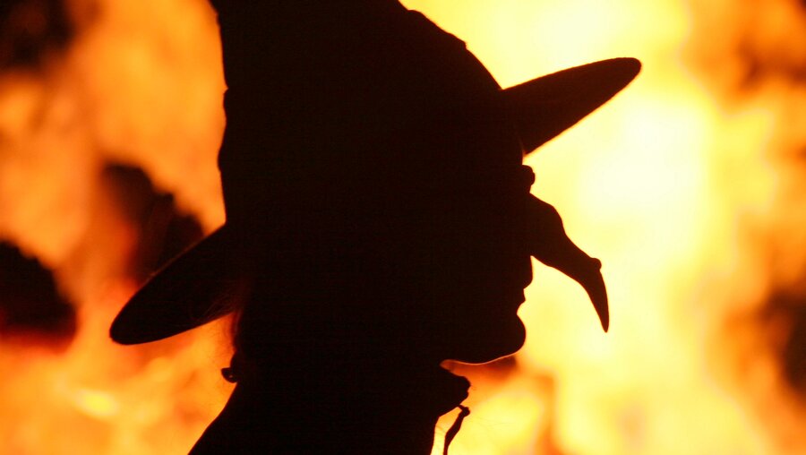 Die Silhouette einer Hexe vor einem Feuer in der Walpurgisnacht. Es gibt Hexen, die mit ihren übernatürlichen Fähigkeiten anderen Schaden zufügen können - daran glaubt eine überraschend große Zahl der Menschen weltweit. / © Matthias Bein (dpa)