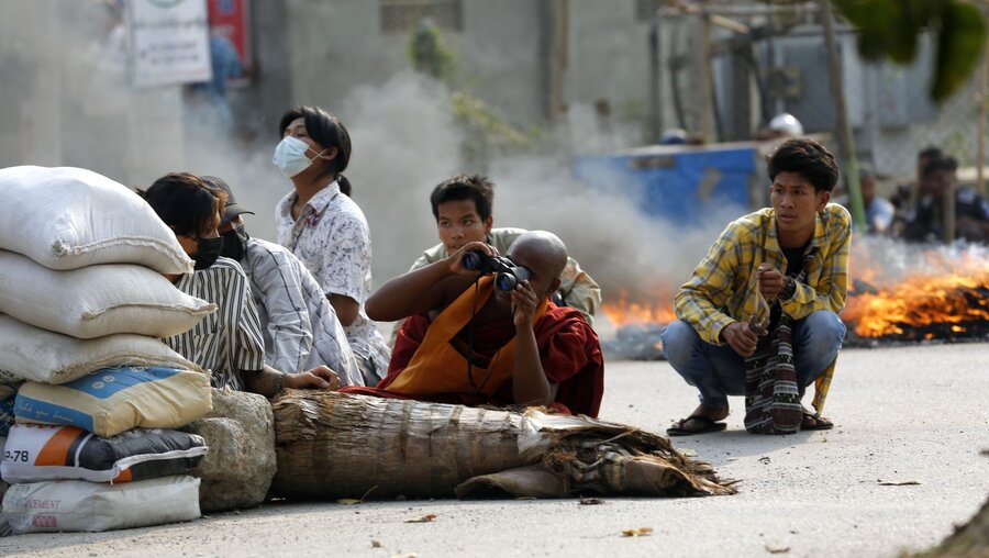 Myanmar, Mandalay: Ein buddhistischer Mönch (M) benutzt ein Fernglas, während er mit anderen Männern hinter einer Straßenabsperrung hockt / © Uncredited/AP (dpa)