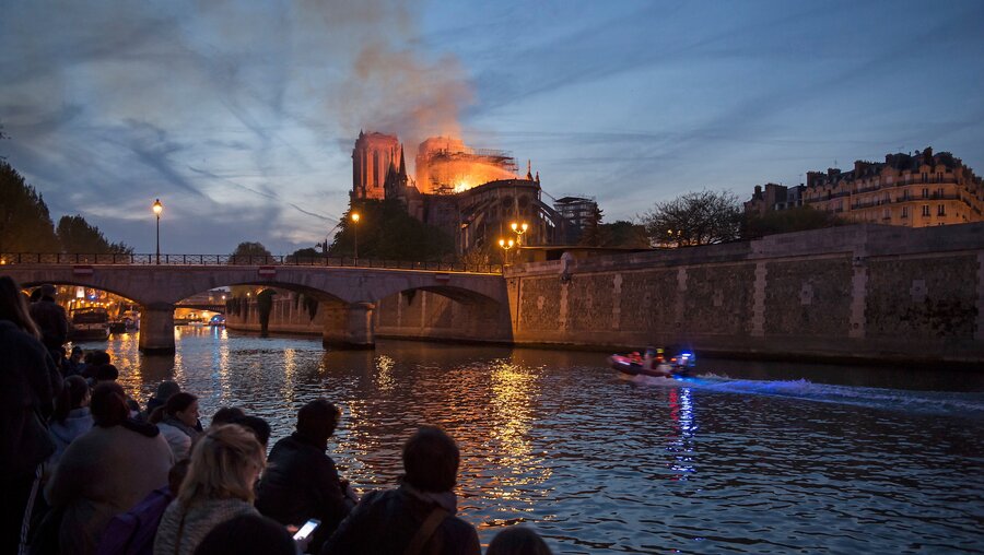 Menschen stehen am Ufer der Seine und blicken auf den Brand der Kathedrale Notre-Dame in Paris am 15. April 2019. / © Corinne Simon (KNA)