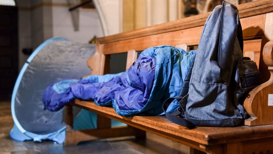 Ein Schlafsack und ein Rucksack liegen auf einer Kirchenbank. Im Hintergrund steht ein Zelt. / © Harald Oppitz (KNA)