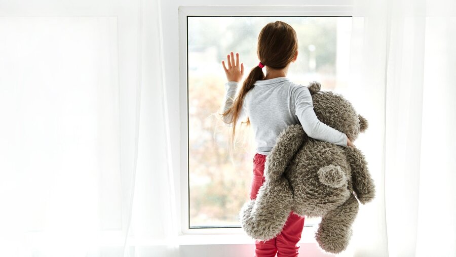 Kind mit Teddybär blickt aus einem Fenster / © PinkCoffee Studio (shutterstock)
