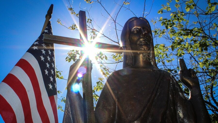 Sonnenstrahlen brechen sich an einem Kreuz, in der Hand einer bronzenen Jesusfigur / © Bradley Birkholz (KNA)