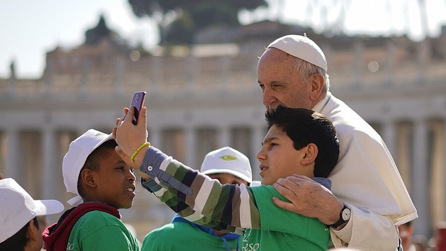Vatikan: Ein Junge macht ein Selfie mit Papst Franziskus, als dieser zu seiner wöchentlichen Audienz auf dem Petersplatz ankommt. / © Andrew Medichini (dpa)