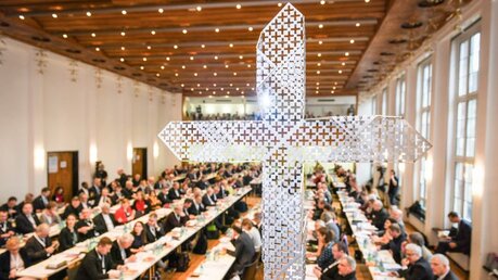 Blick durch ein gelochtes Metallkreuz auf die Teilnehmer an langen Tischen bei den Beratungen der Synodalversammlung / © Harald Oppitz (KNA)