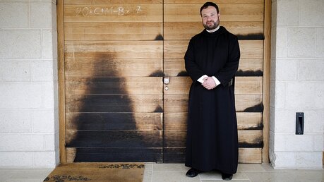 Pater Nikodemus Schnabel vor einer Tür zum Atrium der Brotvermehrungskirche, die noch Brandspuren aufweist / © Corinna Kern (dpa)