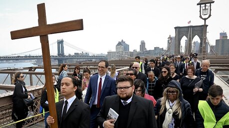 Joshua Layugan (l) trägt ein Kreuz, während er und andere Gläubige bei einer Kreuzwegprozession über die Brooklyn Bridge gehen / © Bebeto Matthews (dpa)