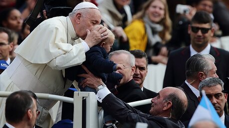 Papst Franziskus küsst den Kopf eines kleinen Kindes, das ihm bei seiner wöchentlichen Generalaudienz entgegen gestreckt wird. / © Evandro Inetti/ZUMA Wire (dpa)