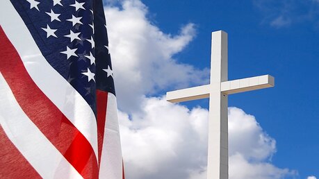 Die Flagge der Vereinigten Staaten von Amerika und ein weißes Kreuz vor blauem Himmel mit weißen Wolken. / © John Panella (shutterstock)