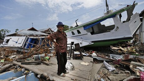 Ein Mann steht inmitten von Trümmern und neben Booten, die von einem Tsunami an Land geschwemmt wurden. / © Dita Alangkara (dpa)
