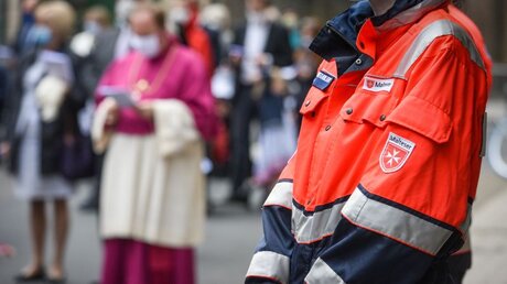 Eine Person mit einer Jacke des Malteser Hilfsdienstes, auf der das Logo mit dem Malteserkreuz angebracht ist, beim Gottesdienst zu Fronleichnam am 11. Juni 2020 in Köln. / © Harald Oppitz (KNA)