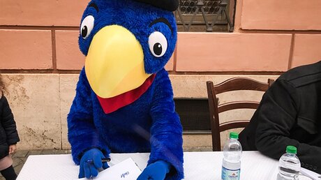 Eine Person, verkleidet als Papagei Globi, eine bekannte Schweizer Kinderbuchfigur, signiert ein Buch / © Stefanie Stahlhofen (KNA)