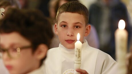 Ein Junge trägt bei seiner Kommunion eine weiße Kommunionalbe und hält eine brennende Kommunionkerze in der Hand.  / © Natalia Gileva (KNA)