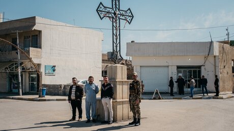 Vier Männer, chaldäische Christen, warten an einem großen Kreuz an einer Straße auf die Durchfahrt von Papst Franziskus nach Erbil am 7. März 2021 in Karamless, Irak. / © Jean-Matthieu Gautier (KNA)