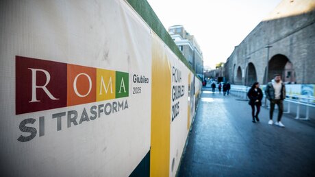 Passanten gehen an einem Bauzaun vorbei mit der Aufschrift "Roma si transforma - Giubileo 2025" (dt. Rom verwandelt sich - Jubiläumsjahr 2025) / © Cristian Gennari/Romano Siciliani (KNA)