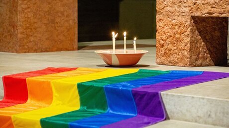 Vier brennende Kerzen stehen in einer Schale neben einer Regenbogenfahne auf den Altarstufen während eines Gottesdienstes zum 20-jährigen Bestehen von Queer-Gottesdiensten in der Kirche Sankt Paul am 13. März 2022 in München. / © Robert Kiderle (KNA)