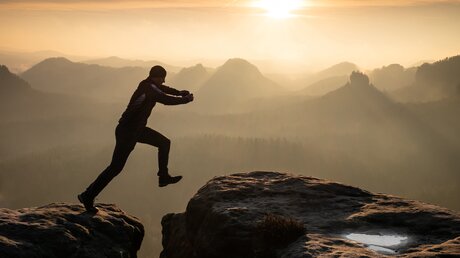 Ein Bergsteiger überspringt bei einer Wanderung einen Gipfelgrat vor einer Bergkulisse bei Sonnenuntergang. (Symbolbild: Glaube, Inspiration, Entdeckergeist) / © rdonar (shutterstock)