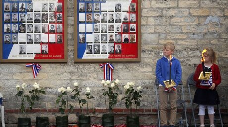 Kinder stehen neben einem Brett mit Bildern von Kriegsopfern bei einer Gedenkfeier / © Francois Mori (dpa)