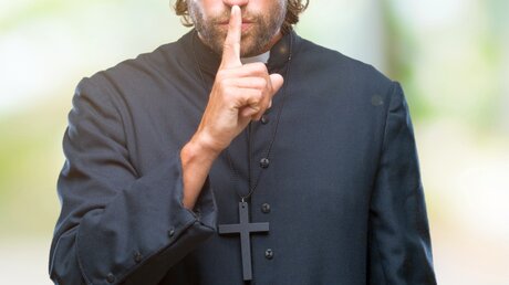 Symbolbild: Pfarrer mit Finger vor dem Mund / © Aaron Amat (shutterstock)