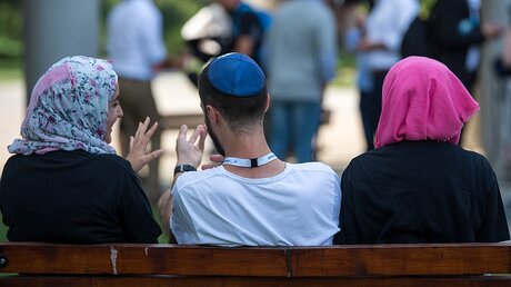 Zwei junge Muslimas mit Kopftuch und ein junger jüdischer Mann mit Kippa unterhalten sich miteinander / © Monika Skolimowska (dpa)