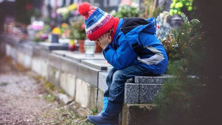 Ein Kind sitzt neben Gräbern auf einem Friedhof / © Tomsickova Tatyana (shutterstock)