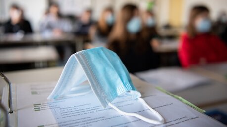Eine Mund- und Nasen-Bedeckung liegt im Unterricht auf Arbeitsmappe / © Matthias Balk (dpa)