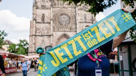 Eine Helferin steht vor der Lorenzkirche und hält einen Schal hoch mit dem Kirchentagsmotto auf dem Deutschen Evangelischen Kirchentag / © Katharina Gebauer (KNA)