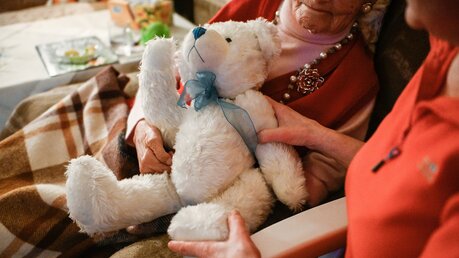 Eine Seniorin sitzt zugedeckt und mit einem Teddybär im Arm zu Hause im Sessel, ihre Pflegerin sitzt neben ihr. / © Julia Steinbrecht (KNA)