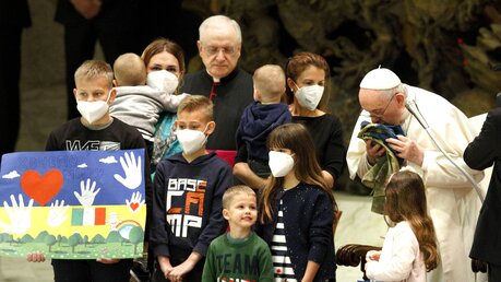 Papst Franziskus steht neben Flüchtlingskindern und -frauen aus der Ukraine und küsst eine ukrainische Nationalflagge, die er aus dem ukrainischen Ort Butscha erhalten hat, bei der Generalaudienz am 6. April 2022 im Vatikan.  / © Paul Haring (KNA)
