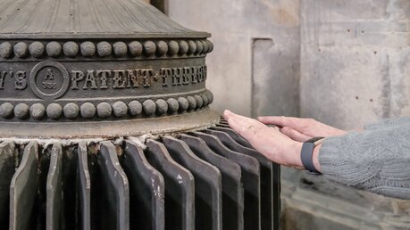 Hände wärmen an einem sehr alten, funktionierenden Dampfkessel in einer Kathedrale in Ely, Cambridgeshire, UK. / © Nick Beer (shutterstock)