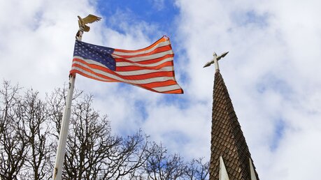 Symbolbild US-Fahne neben einem Kirchturm / © Bobkeenan Photography (shutterstock)