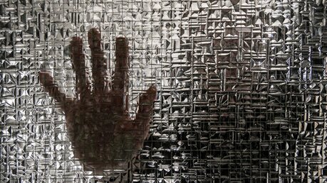 Die Umrisse einer Hand, sie sich auf eine Glasscheibe stützt, dahinter die unscharfe Silhouette einer Person. / © Harald Oppitz (KNA)