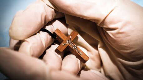 Mann mit medizinischen Handschuhen hält ein Kreuz / © elena_prosvirova (shutterstock)