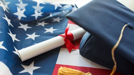 Symbolbild: Bachelor-Hut und Diplom unter amerikanischer Flagge / © Syda Productions (shutterstock)