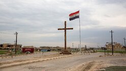 Ein Kreuz und eine irakische Flagge an einer Straße am Ortseingang von Karakosch. / © Uygar Onder Simsek (KNA)